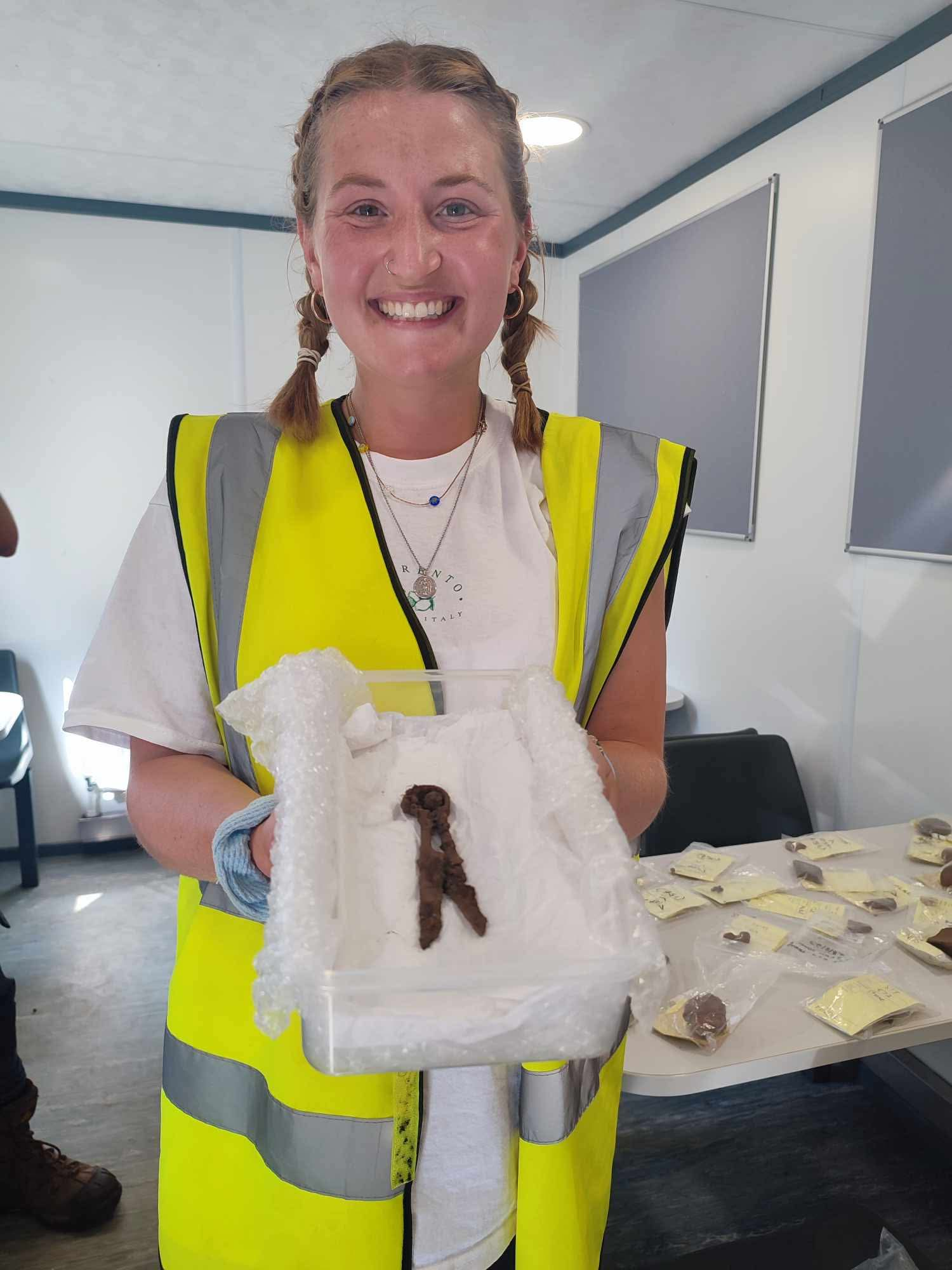 Emily's figure takes up the majority of the frame - standing in the centre from hips up. She wears a white t-shirt and high vis vest. She smiles directly at the camera and hair is in French plaits. In her hands she is holding a Tupperware box with iron shears in them, which are rusted, from excavation