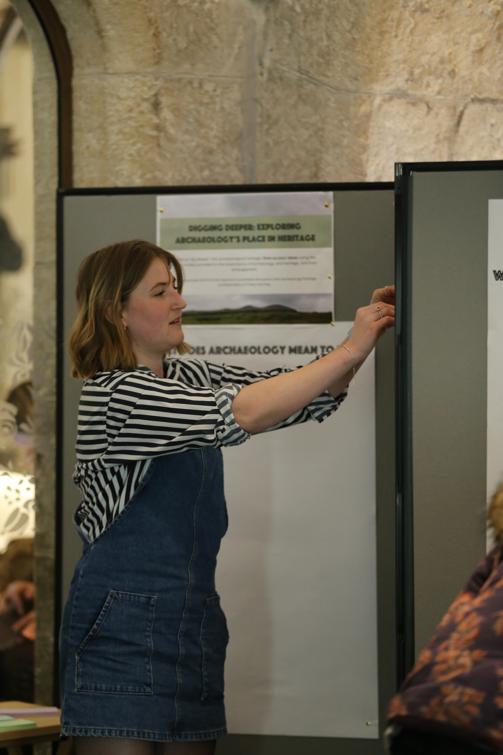 Emily stands to the left of the image in a blue denim pinafore dress and navy striped shirt. Her hands are in front of her pining things to a notice board. Behind her are posters about Archaeology