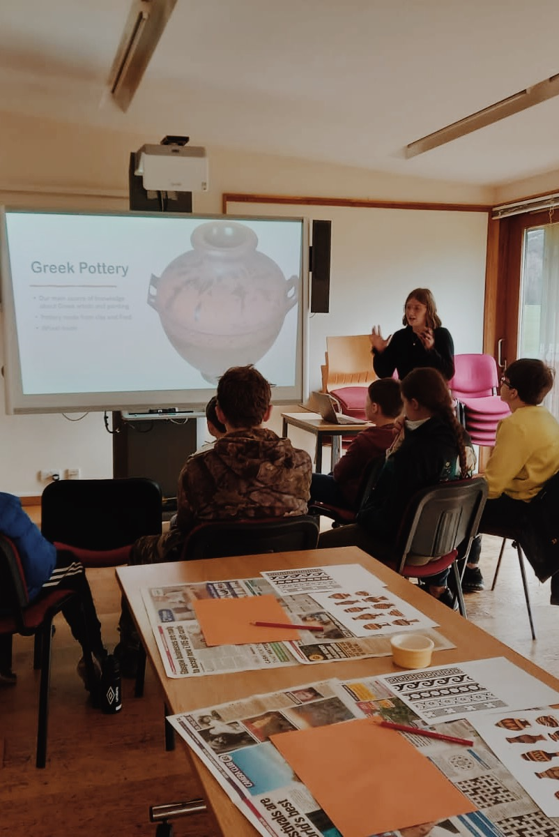 The bottom third of the image is a table with photos of greek vases and arts and crafts activities. Behind the table are the backs of children sitting looking at a screen which has 'Greek Pottery' on it and a vase image. Emily stands next to the screen with both hands up as she gestures at the image whilst talking
